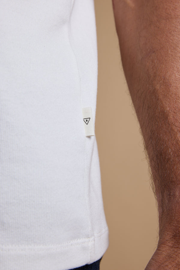 Close-up of a person wearing the Barkers Langwood Tee in white organic cotton, featuring a small white tag with a black triangle logo on the side seam. The relaxed fit stands out against a beige background.