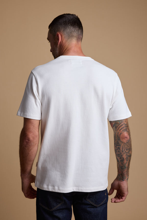 A man with short dark hair, a trimmed beard, and a tattoo sleeve on his right arm stands with his back to the camera, wearing the Barkers Langwood Tee and dark jeans against a beige background.