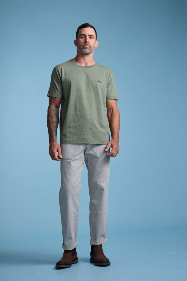 A man with short dark hair and visible arm tattoos stands against a plain light blue background, wearing the Barkers Bayside Emb Tee, light gray pants, and brown shoes, with a neutral facial expression.