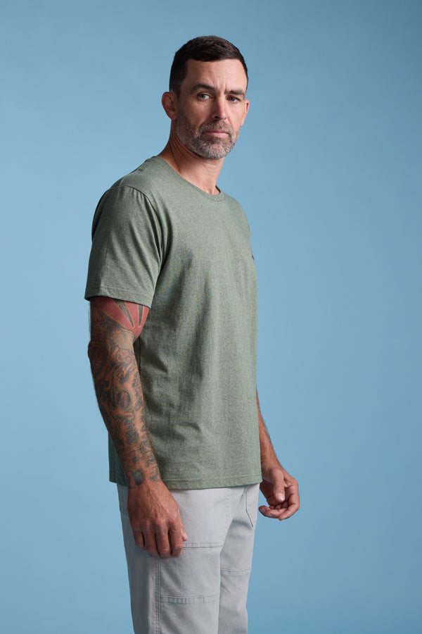 A man with short dark hair and a beard, in the Barkers Bayside Emb Tee (green organic cotton) and light gray pants, stands against a blue background. Tattoos are visible on his left arm as he looks slightly to the side.