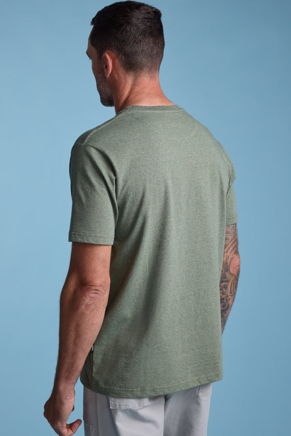 A man with short dark hair, seen from behind, wears the Barkers Bayside Emb Tee in light green and light gray shorts. A large tattoo is visible on his left arm. The background is a solid light blue.