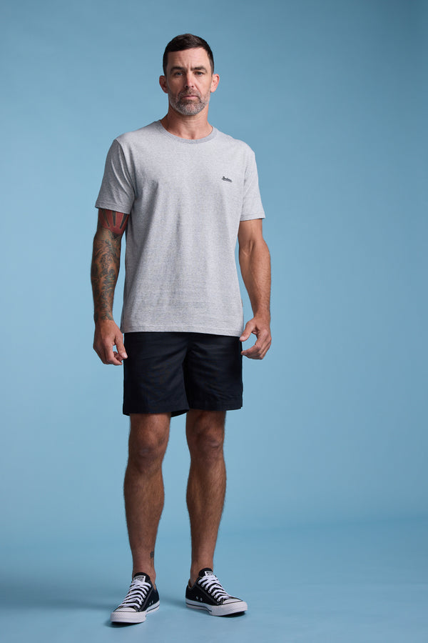 A man with short hair and tattoos on his left arm stands against a light blue background, wearing the Barkers Bayside Emb Tee, black shorts, and black-and-white sneakers.