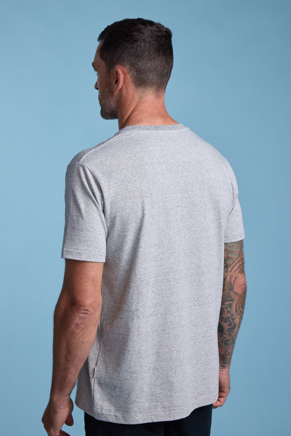 A man with short dark hair and a tattooed right arm is seen from behind, wearing the Barkers Bayside Emb Tee in light gray and dark pants, standing against a plain light blue background.
