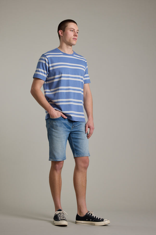 A man stands against a plain background, wearing a blue and white striped t-shirt, Barkers Rover Denim Shorts made from regenerative cotton, and black sneakers. His left hand is in his pocket as he looks slightly to the side.