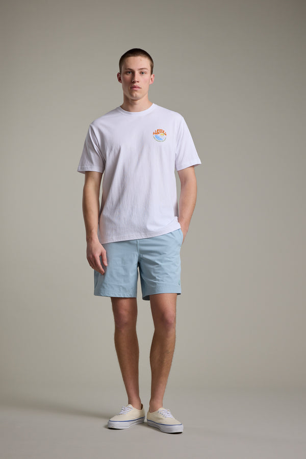 A person stands against a plain background wearing a white T-shirt with a small colorful graphic, Barkers Davis Weekend Short in light blue recycled polyester, and white sneakers. Their hands are at their sides and their expression is neutral.