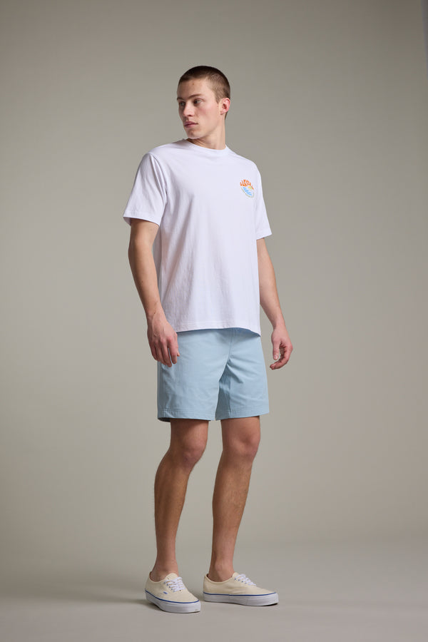 A man stands against a plain background wearing a white T-shirt with a small chest logo, Barkers Davis Weekend Short in light blue, and beige sneakers, looking to his left.