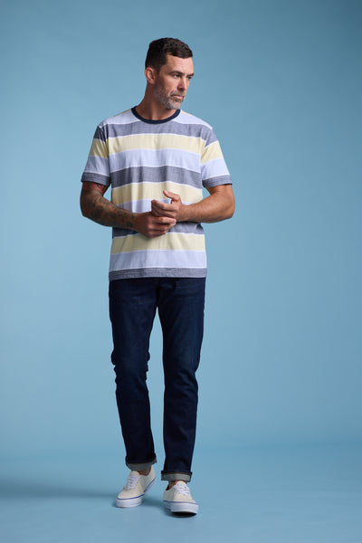 A man with short dark hair and a beard, wearing the Barkers Asbrooke Stripe Tee in pastel shades, dark jeans, and white sneakers, stands against a light blue background looking to his left with his hands clasped.