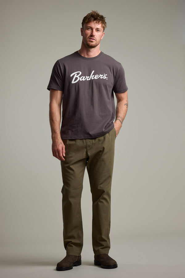 A man with short light brown hair poses against a plain background, wearing a Barkers Finely Tuned Print Tee in grey, paired with olive green pants and brown suede shoes. He stands relaxed with one hand by his side.
