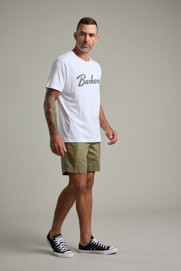 A man with short hair and a beard poses in a studio, wearing the Barkers Finely Tuned Print Tee in white, khaki shorts, and black sneakers. He displays tattoos on his arms while standing against a plain light background.