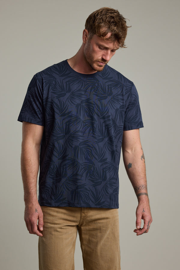 A man with short brown hair and arm tattoos wears the Barkers Palm Leaves Print Tee and beige pants, looking down with a neutral expression against a plain background.