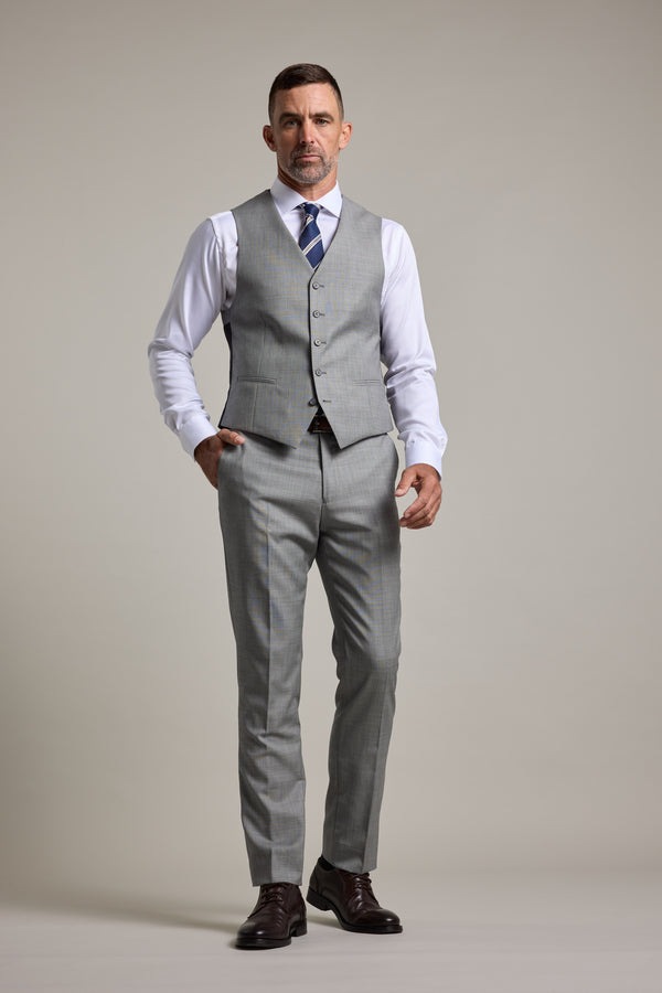 A man with short hair and a neutral expression stands against a plain background wearing the Barkers Henry Waistcoat in premium wool light grey as part of a three-piece suit, paired with a white shirt, blue striped tie, and dark brown dress shoes.