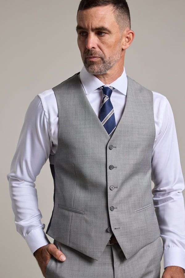 A man with short dark hair and a beard models the Barkers Henry Waistcoat over a white dress shirt and navy striped tie, standing with one hand in his pocket and looking to the side against a plain backdrop.