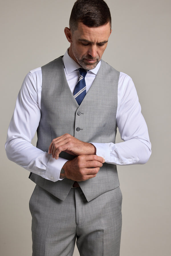 A man in the Barkers Henry Waistcoat and matching pants adjusts the cuff of his white dress shirt. He looks down, wearing a striped tie, against a plain light background.