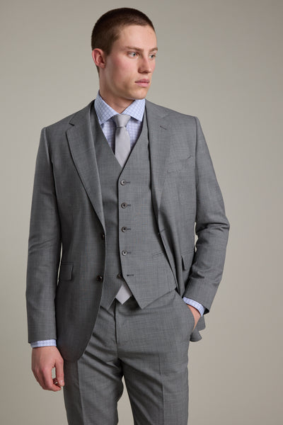 A man stands with one hand in his pocket, looking left, wearing the Barkers Henderson Waistcoat—a refined grey houndstooth piece—paired with a light gray three-piece suit, matching tie, and patterned dress shirt against a plain background.
