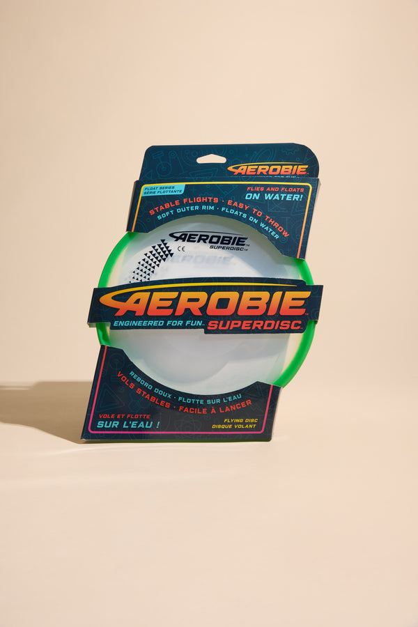 A packaged Aerobie Superdisc by Other, featuring a green rim and clear center, stands upright against a beige background. The packaging notes it flies and floats, is easy to throw, and designed for frisbee fun.