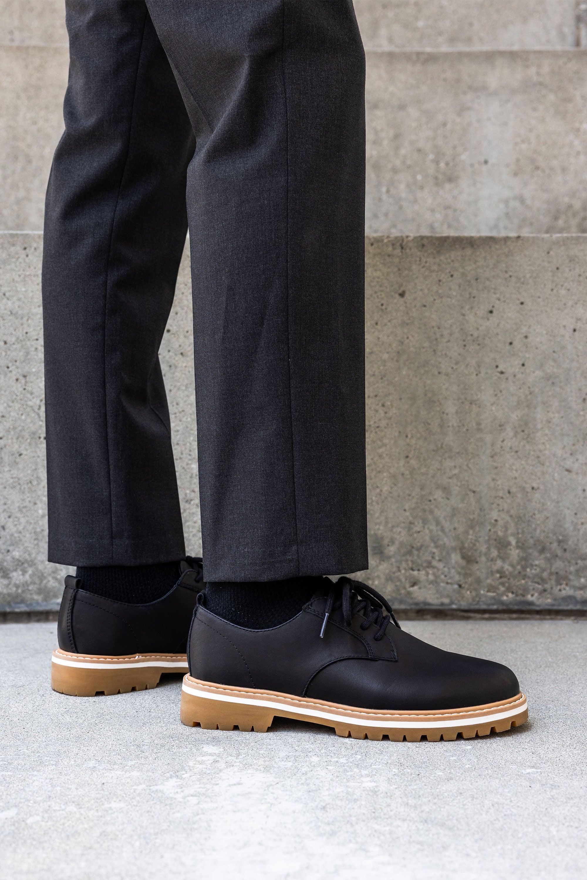 Someone wearing dark dress pants and Clae’s Logan black derby sneakers with light brown rubber soles and a recycled PU foam insole stands on concrete before a staircase.