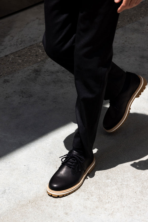 A person wearing black dress pants and CLAE Logan premium leather footwear by Clae with light brown soles is walking on a sunlit concrete surface.