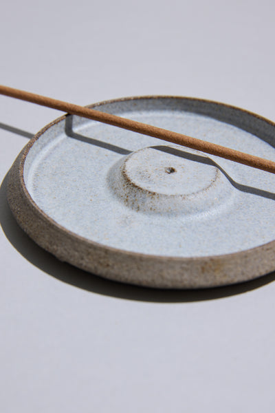 The Studio Foon Incense Dish by Other is a light-colored, handcrafted ceramic piece from New Zealand with a round base, shown holding a single stick of brown incense on a plain, light background.