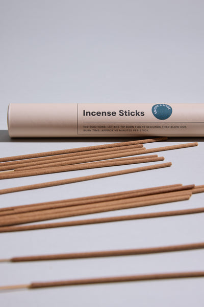 A beige tube labeled "EoE Smoke And Musk Incense Sticks" from Earl of East rests on a white surface, with several brown incense sticks laid out in front. The label features brief instructions for use.