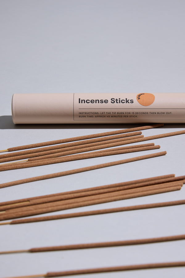 A cylindrical beige Earl of East container labeled "EoE Strand Incense Sticks" rests on a light gray surface, with several birchwood-scented incense sticks arranged in front.