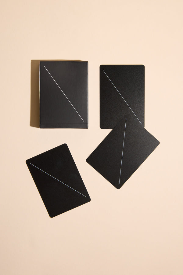 Four Minim Playing Cards by Other, featuring thin diagonal lines, are arranged on a beige surface—three face down and one in a black box.