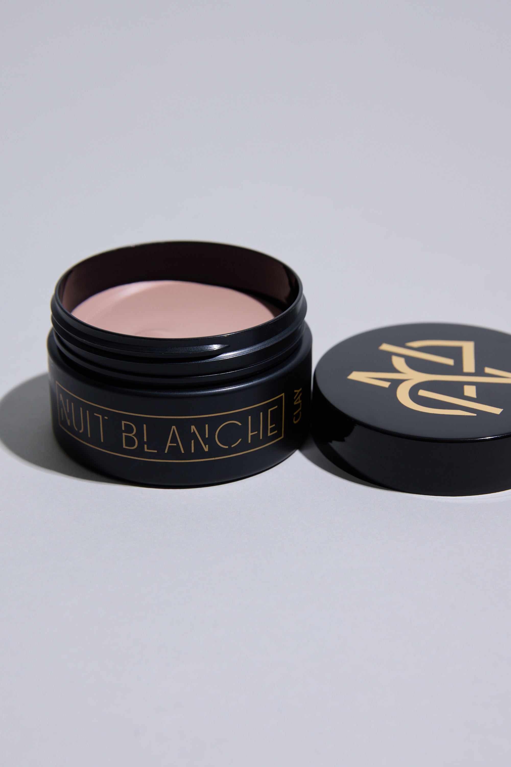 A black jar labeled "Nuit Blanche Clay" by Other sits open on a light gray surface, revealing pale pink matte hair clay for long-lasting hold. The matching black lid with gold accents rests beside the jar.