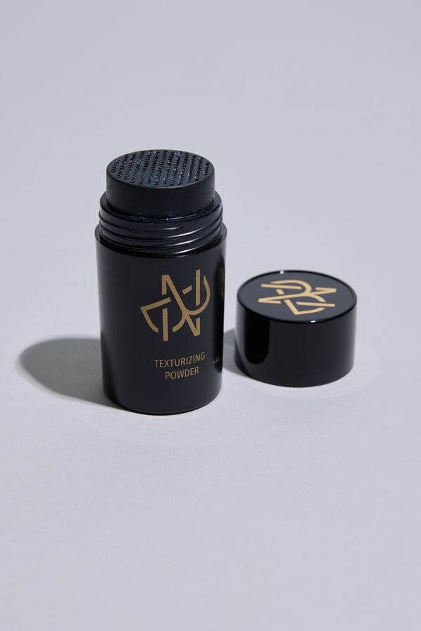 An open black container of Other Nuit Blanche Powder with a gold logo sits on a light gray surface. The cap, also with the gold logo, is to the right. This styling powder boosts hair volume and adds a natural matte finish.