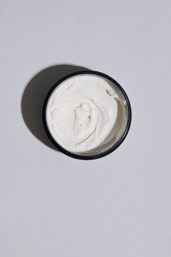 A round black container of Other Nuit Blanche Paste, filled with creamy white hair paste, sits on a plain light gray surface and casts a soft shadow to the left.