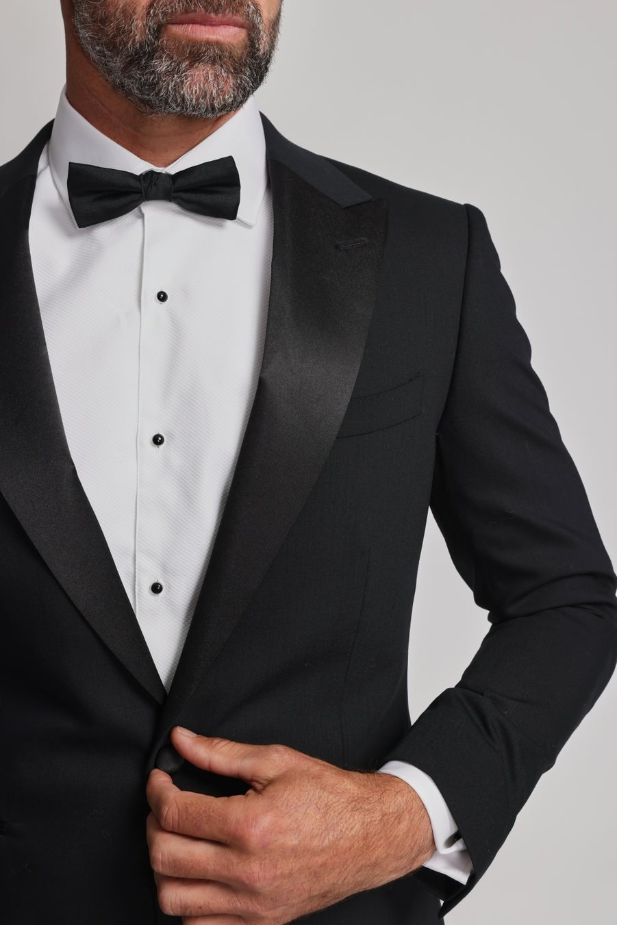 A man in a black tuxedo and white shirt adjusts his jacket, showcasing the Barkers Black Bow Tie with an adjustable neck strap. The image is cropped to display only his torso and lower face.