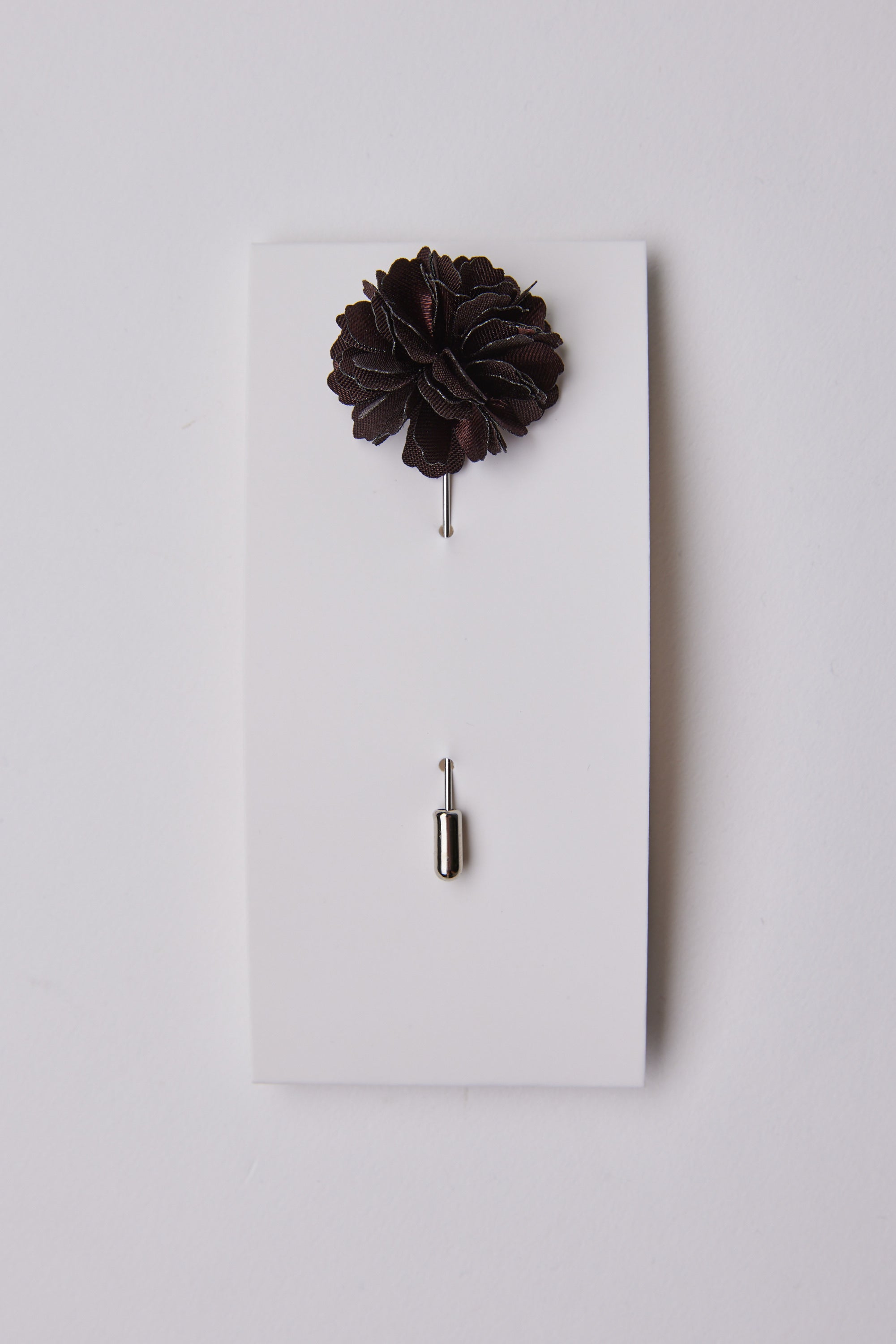 The Parisian Fraser Flower Lapel Pin features a dark floral design with a silver stem, elegantly presented on a white rectangular card. The cap sits below the pin—an ideal accessory for weddings or formal occasions.