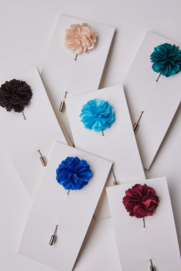 The Parisian Fraser Flower Lapel Pin comes in peach, teal, brown, light blue, deep blue, burgundy, and red—ideal suit accessories for formal events. Displayed on white cards against a crisp white background.