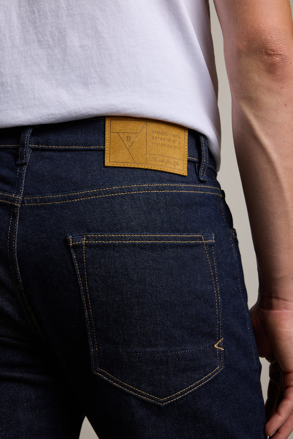 A person shown from behind wears Barkers Rambler Denim Slim Jeans in dark blue with yellow stitching and a tan leather patch on the waistband, paired with a white T-shirt.
