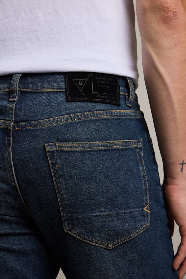 Someone wearing Barkers Rambler Denim Slim Jeans and a white t-shirt is shown from the back. Their right wrist, sporting a small cross tattoo, is visible. The blue jeans feature a black Barkers patch above the back pocket.