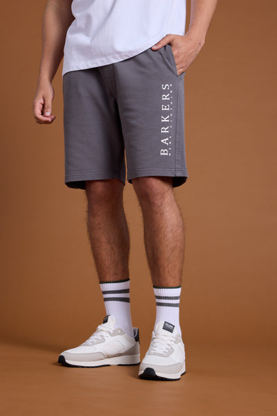 A person wears Barkers Organic Classic Print Track Shorts, paired with a white T-shirt, white crew socks with black stripes, and white sneakers. Only their lower body is shown against a brown background.
