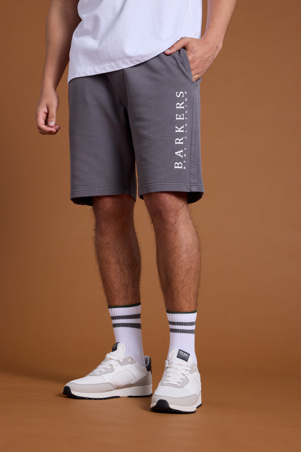 A person wears Barkers Organic Classic Print Track Shorts, paired with a white T-shirt, white crew socks with black stripes, and white sneakers. Only their lower body is shown against a brown background.