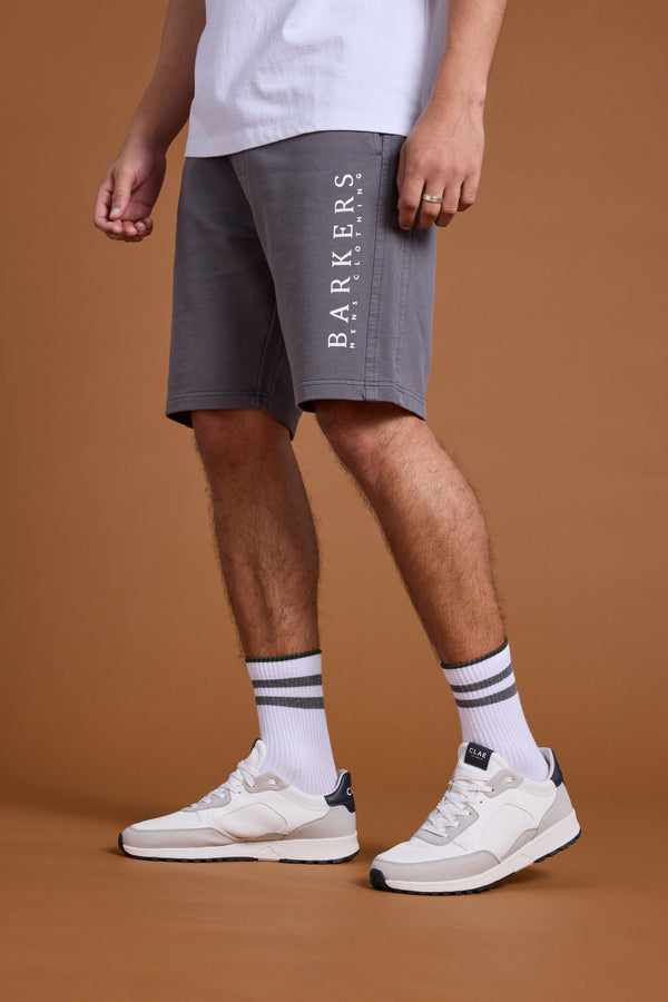 A person models Barkers Organic Classic Print Track Shorts in gray with a white tee, striped white socks, and white sneakers against a plain brown backdrop. Only the lower half is shown.