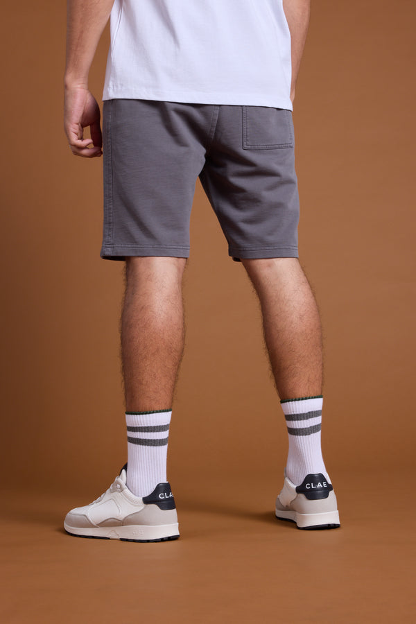 A person stands with their back to the camera, wearing Barkers Organic Classic Print Track Shorts in gray, a white t-shirt, white sneakers, and white socks with gray stripes, set against a brown background.