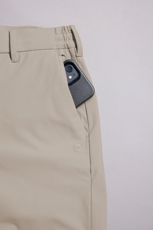 A close-up of WearEver Switch Shorts in beige comfort stretch, showing a smartphone partially tucked into the right pocket, with belt loops and a subtle embroidered WearEver logo near the pocket.