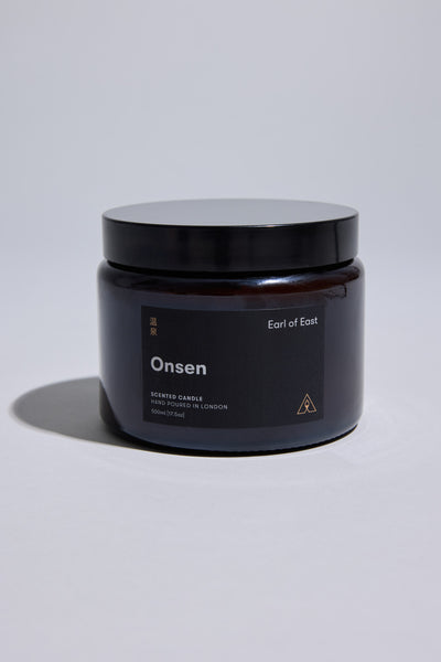 A dark amber glass jar with a black lid displays the Earl of East EoE Onsen Candle 500ml, hand-poured in London and inspired by Japanese hot springs. The product is set against a plain light background.