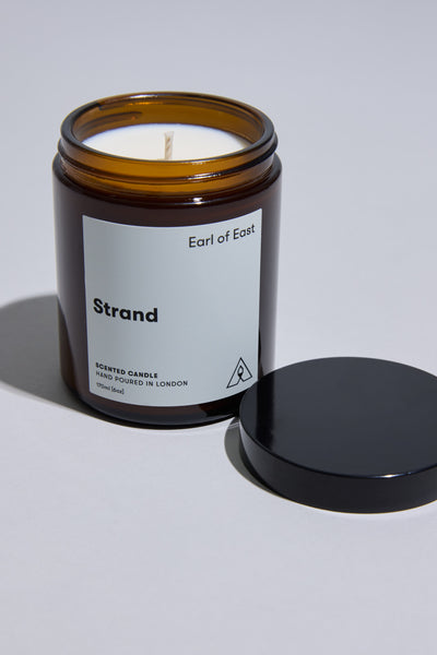 A 170ml EoE Strand Candle by Earl of East, in a brown glass jar with a white label, sits on a light gray surface. The black lid is off, placed next to the scented candle, ready to release mandarin notes.