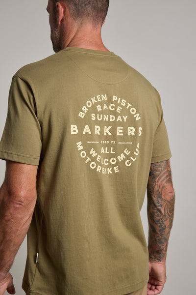 A man with a tattooed arm, facing away, wears the Barkers Sunday Club Print Tee in olive-green. Made from organic cotton, it features white "Broken Piston Race Sunday, Barkers, All Welcome, Motorbike Club" text on the back.