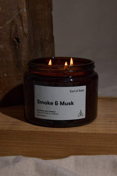 A lit 500ml EoE Smoke And Musk Candle by Earl of East, made from soy wax and patchouli, glows on a wooden surface against a rustic backdrop under soft, warm light.