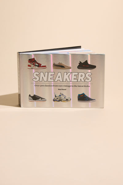 The hardcover book "Sneakers" by Neal Heard (Other) is shown against a beige background, featuring iconic sneakers on the cover—a must-have anthology for sneaker enthusiasts, highlighting over 300 classics from vintage to modern styles.