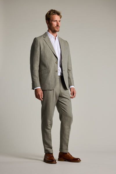 A man stands against a plain background in a light gray linen-cotton blend suit, white dress shirt, and brown shoes, looking off to the side. He showcases refined summer style with Barkers' Paxton Linen Pant.