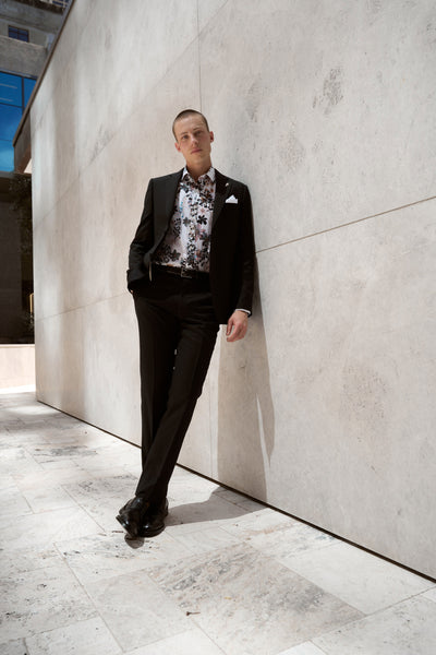 Wearing the Barkers Apothecary AOP Business Shirt in classic black, crafted from responsibly sourced cotton, a person leans casually against a light stone wall outdoors with hands in pockets as sunlight casts soft shadows nearby.