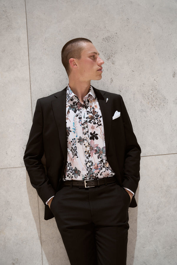 A man in a black suit and Barkers Apothecary AOP Business Shirt stands against a light stone wall with his hands in his pockets, looking to the side.