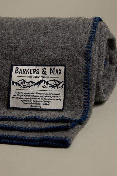 A grey recycled Wool Take Back Blanket by Other with blue trim is rolled up, featuring a white label reading “Other Made in New Zealand” with mountain graphics and product details in smaller text.