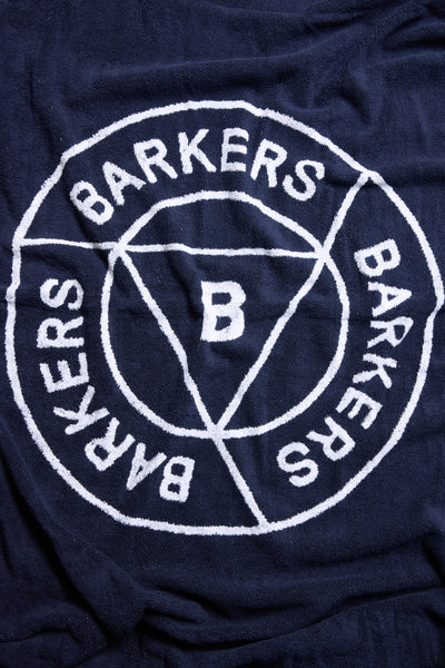 The Barkers Beach Towel is navy blue, featuring a white circular Barkers logo with "BARKERS" repeated three times and a central B, intersected by lines forming a triangle. Made from organic cotton for lasting softness and durability.