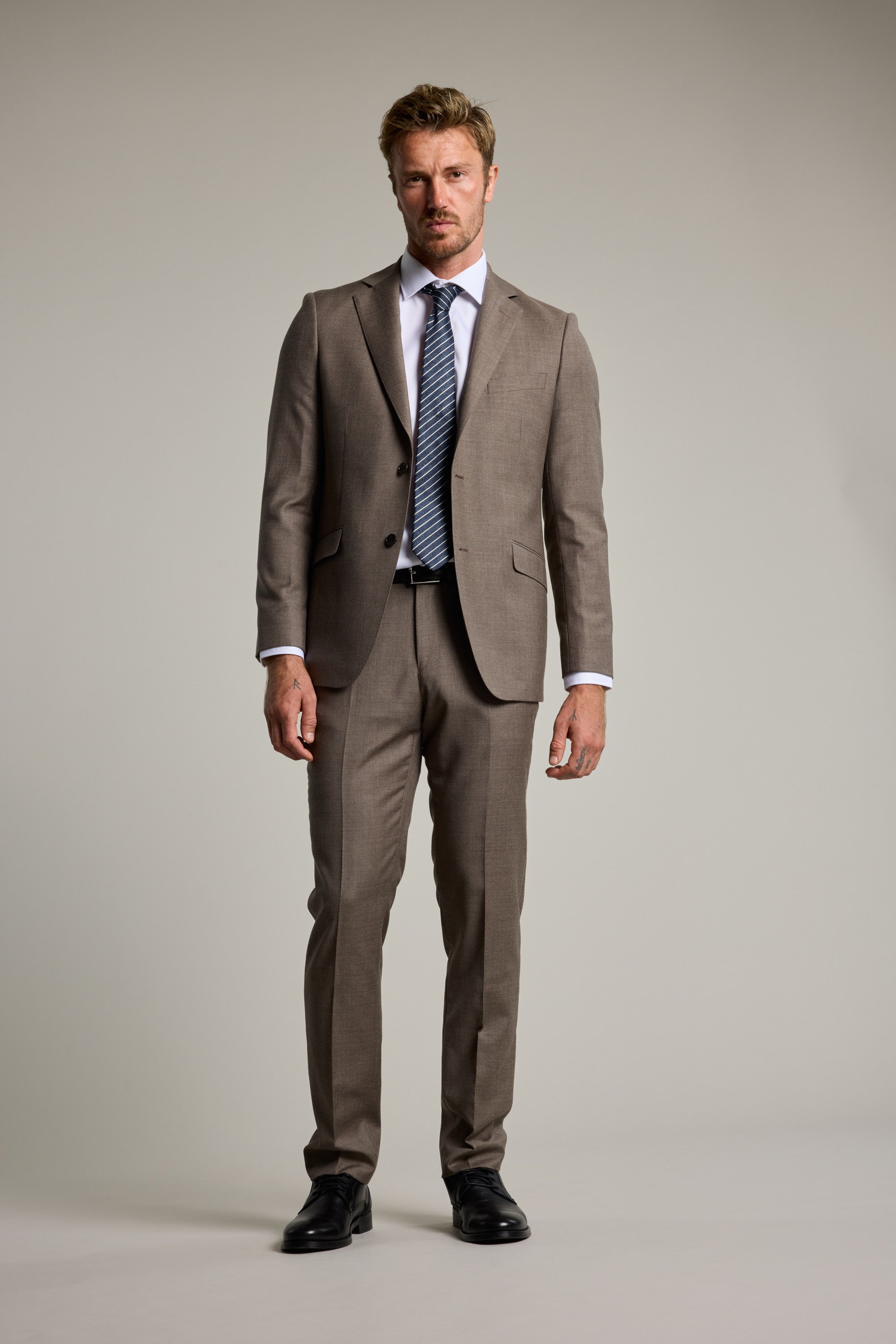A man stands facing forward in a Barkers Westport Melange Suit Jacket, paired with a white dress shirt, striped tie, and black dress shoes against a plain light gray background.