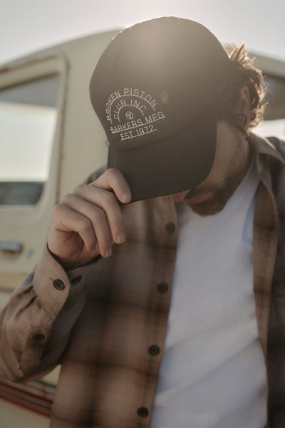 A man outdoors by a cream vehicle tilts his black Barkers Piston Club Trucker Cap with an embroidered logo, partially covering his face as sunlight highlights his plaid shirt and white t-shirt.